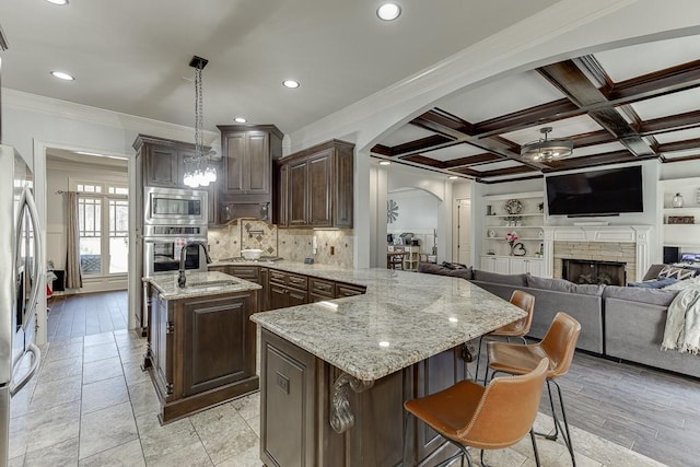 kitchen with stainless steel appliances, light stone counters, pendant lighting, coffered ceiling, and a kitchen island with sink