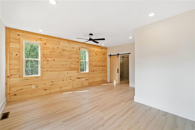 empty room with ceiling fan, a barn door, wooden walls, and light hardwood / wood-style flooring