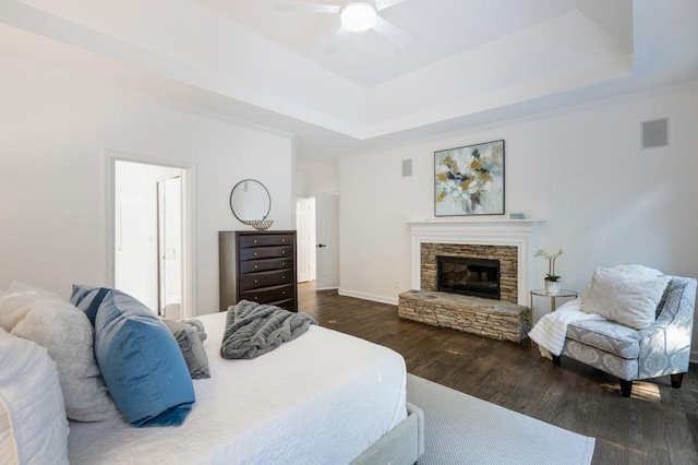 bedroom with a stone fireplace, a tray ceiling, dark wood-type flooring, and ceiling fan