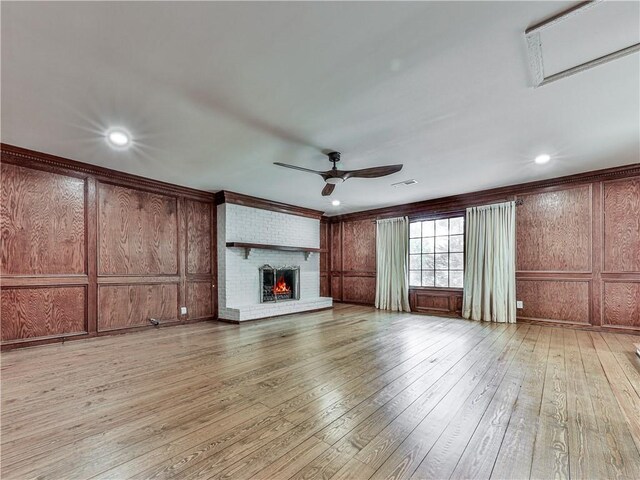 unfurnished living room featuring ceiling fan, light wood-type flooring, wooden walls, and a brick fireplace