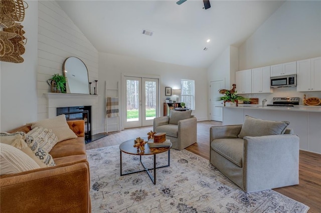 living area featuring light wood finished floors, visible vents, a glass covered fireplace, ceiling fan, and high vaulted ceiling