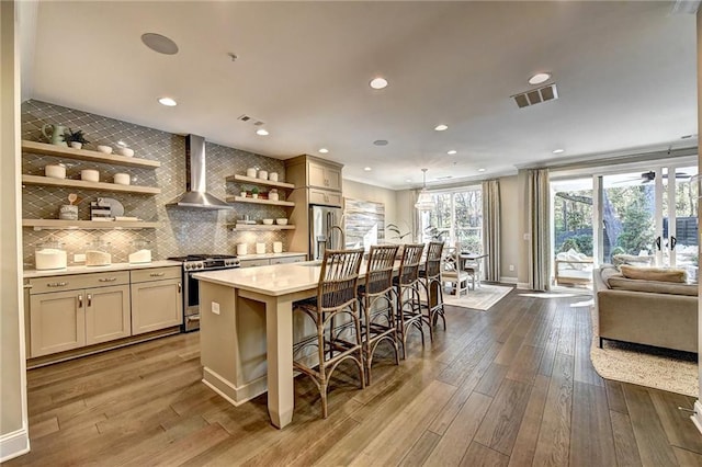 kitchen with wall chimney exhaust hood, stainless steel appliances, hardwood / wood-style floors, a breakfast bar area, and a center island with sink