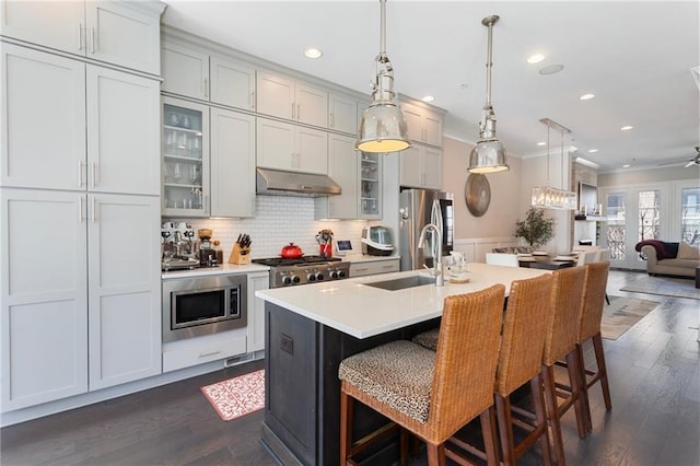 kitchen with stainless steel appliances, under cabinet range hood, ornamental molding, dark wood-style flooring, and a sink
