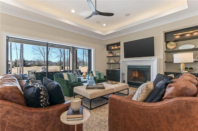 living room with built in features, a tray ceiling, recessed lighting, ceiling fan, and a lit fireplace