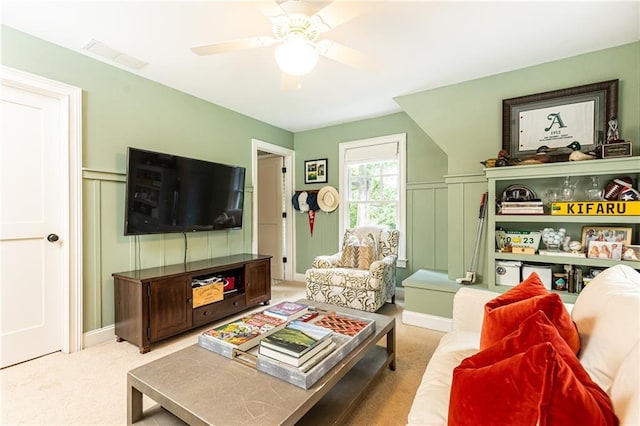 carpeted living area featuring a decorative wall, a ceiling fan, and wainscoting