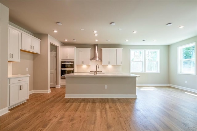 kitchen with light wood finished floors, baseboards, wall chimney exhaust hood, a kitchen island with sink, and a sink