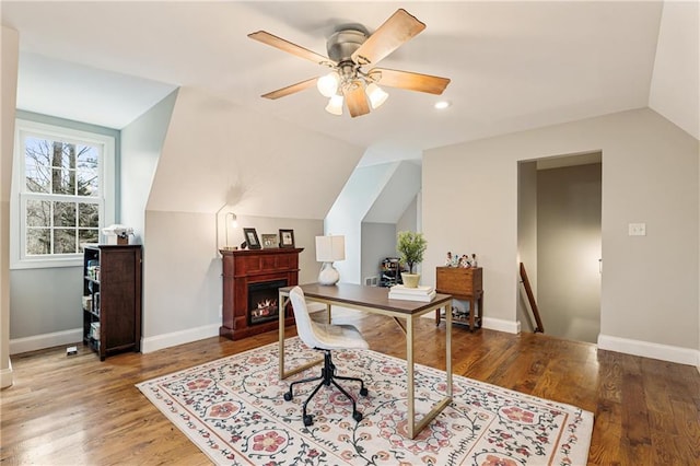 office space with a warm lit fireplace, baseboards, a ceiling fan, lofted ceiling, and wood finished floors