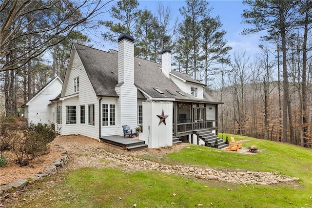 rear view of house with a shingled roof, a fire pit, a lawn, a sunroom, and a wooden deck