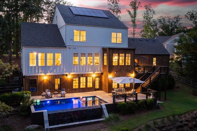 back of property at dusk with solar panels, roof with shingles, a wooden deck, an outdoor pool, and a patio area