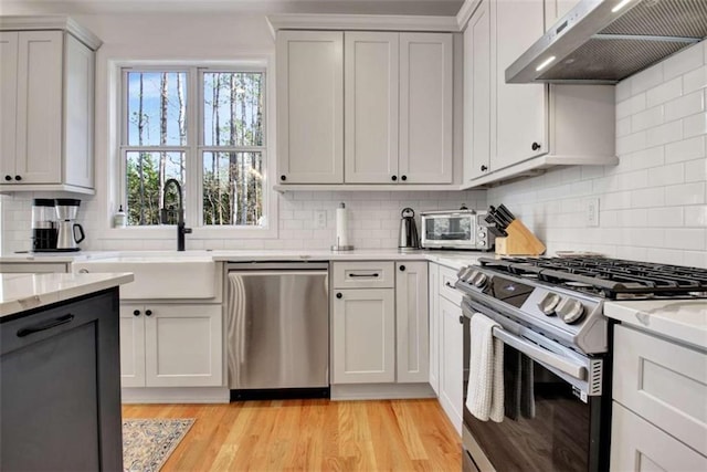 kitchen with appliances with stainless steel finishes, white cabinetry, sink, exhaust hood, and light hardwood / wood-style flooring