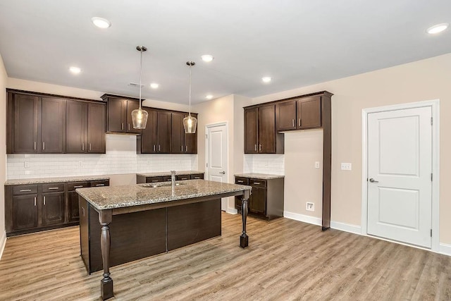 kitchen with light stone counters, backsplash, decorative light fixtures, a center island with sink, and light wood-type flooring