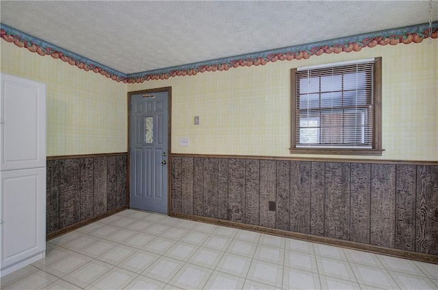 spare room featuring wallpapered walls, baseboards, a wainscoted wall, a textured ceiling, and light floors