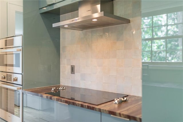 kitchen with butcher block counters, tasteful backsplash, black electric stovetop, stainless steel double oven, and wall chimney exhaust hood