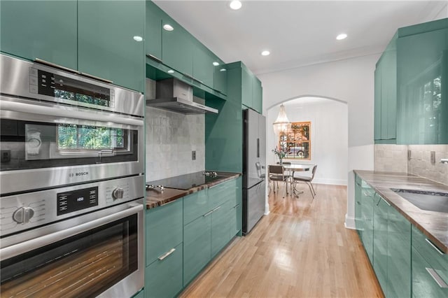 kitchen with light hardwood / wood-style floors, green cabinetry, wall chimney exhaust hood, appliances with stainless steel finishes, and backsplash