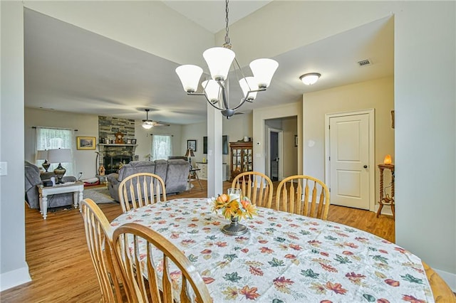 dining room featuring ceiling fan with notable chandelier, a healthy amount of sunlight, a stone fireplace, and light hardwood / wood-style flooring