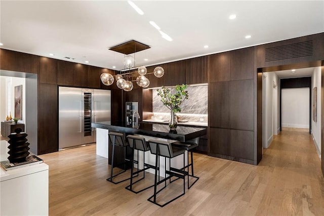 kitchen featuring pendant lighting, a center island, stainless steel built in fridge, light wood-type flooring, and dark brown cabinets