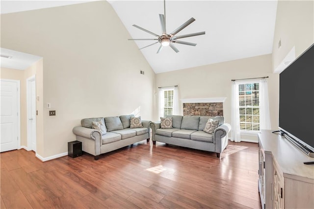 living area featuring a ceiling fan, wood finished floors, baseboards, high vaulted ceiling, and a stone fireplace