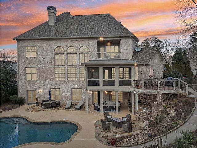 back of house at dusk with an outdoor living space, an outdoor pool, a balcony, a chimney, and a patio area