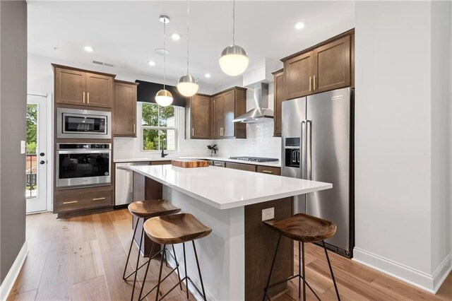 kitchen featuring stainless steel appliances, light wood-style floors, a kitchen breakfast bar, wall chimney exhaust hood, and tasteful backsplash