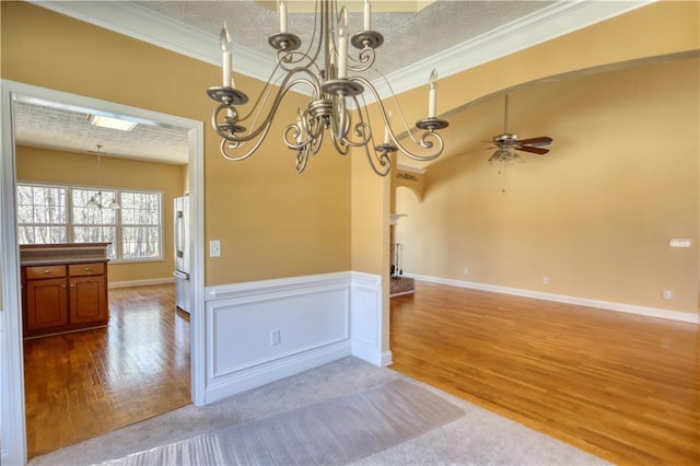 spare room with arched walkways, ceiling fan with notable chandelier, a wainscoted wall, wood finished floors, and crown molding