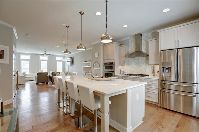 kitchen with light countertops, appliances with stainless steel finishes, decorative backsplash, wall chimney exhaust hood, and light wood finished floors