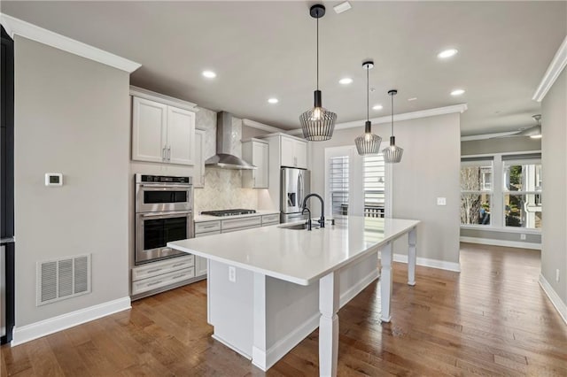 kitchen with a center island with sink, visible vents, appliances with stainless steel finishes, a sink, and wall chimney range hood