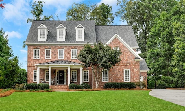 colonial-style house featuring a shingled roof, covered porch, brick siding, and a front lawn