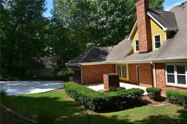 view of side of home featuring a lawn, a patio, roof with shingles, brick siding, and a chimney