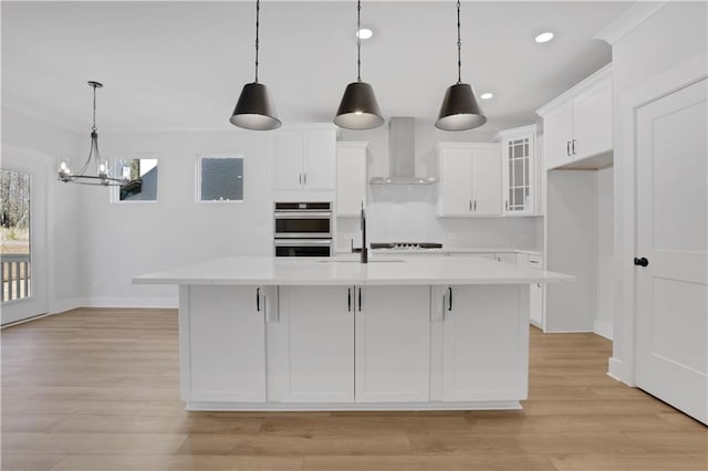 kitchen featuring an island with sink, glass insert cabinets, decorative light fixtures, light countertops, and wall chimney range hood