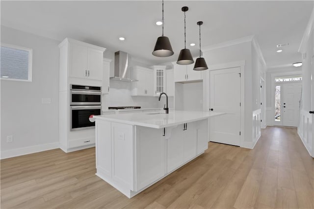 kitchen with double oven, glass insert cabinets, light countertops, wall chimney range hood, and a center island with sink