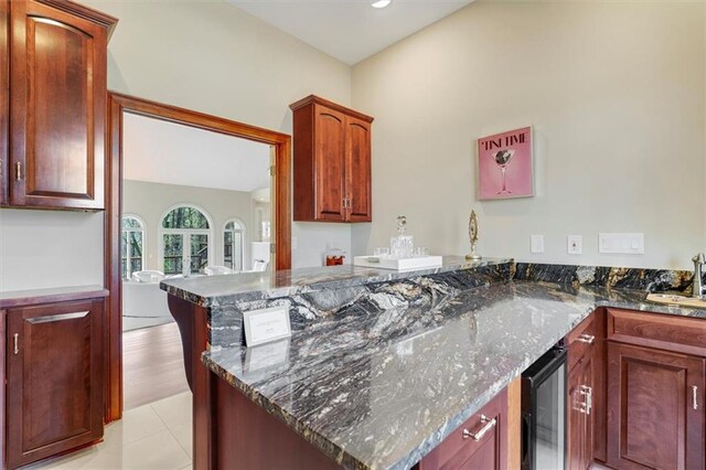 kitchen with light tile patterned floors, a sink, dark stone countertops, beverage cooler, and a peninsula