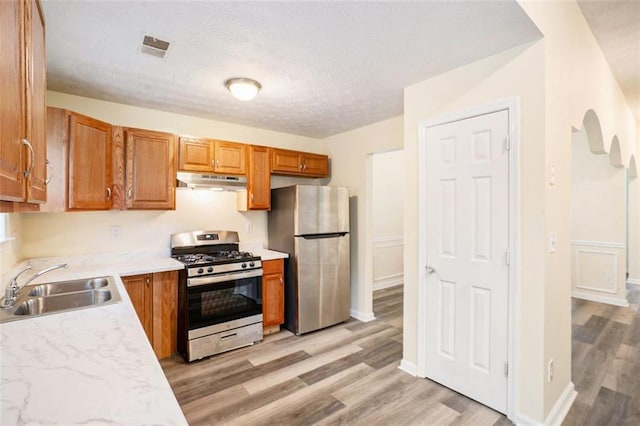kitchen featuring a sink, brown cabinetry, appliances with stainless steel finishes, under cabinet range hood, and light wood-style floors
