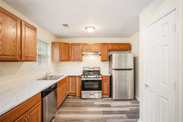 kitchen featuring a sink, stainless steel appliances, brown cabinets, under cabinet range hood, and light wood finished floors