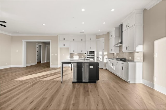 kitchen featuring white cabinetry, an island with sink, built in microwave, ornamental molding, and wall chimney exhaust hood