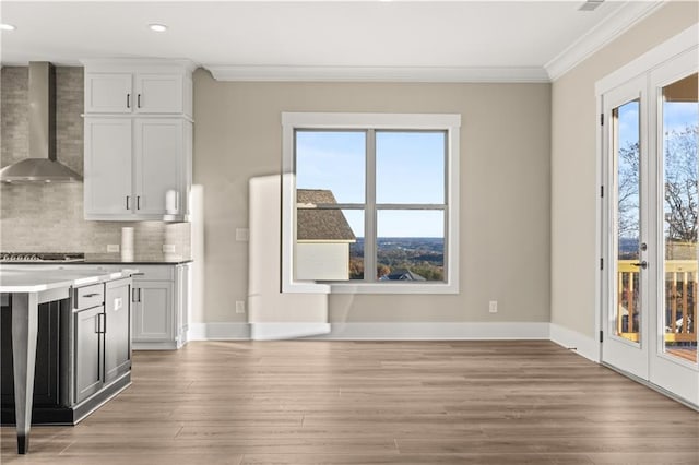 kitchen featuring white cabinetry, light hardwood / wood-style floors, tasteful backsplash, wall chimney range hood, and crown molding