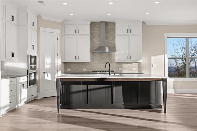 kitchen featuring white cabinets, a kitchen island with sink, wall chimney exhaust hood, and crown molding