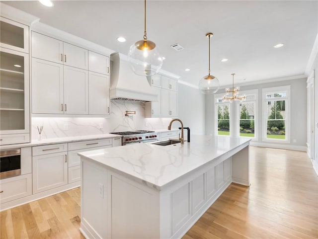 kitchen featuring premium range hood, a kitchen island with sink, sink, and white cabinets