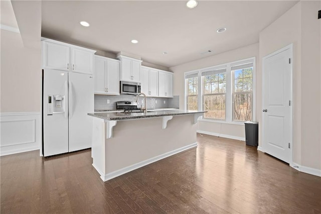 kitchen with a center island with sink, stainless steel appliances, white cabinets, dark stone countertops, and a kitchen bar