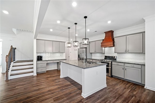 kitchen with appliances with stainless steel finishes, premium range hood, dark stone counters, a center island with sink, and decorative light fixtures