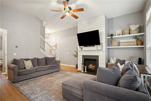 living area featuring stairway, a large fireplace, a ceiling fan, and wood finished floors