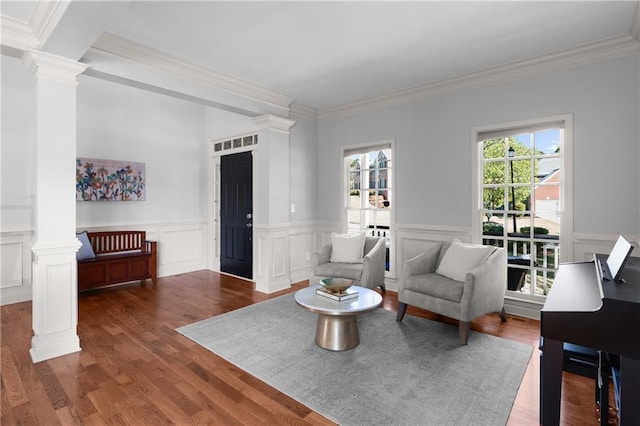 living area featuring ornate columns, crown molding, wood finished floors, and a wainscoted wall