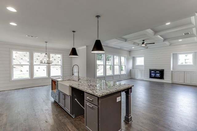 kitchen with coffered ceiling, an island with sink, light stone counters, open floor plan, and a sink