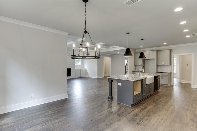 kitchen with light stone counters, a kitchen island with sink, crown molding, gray cabinetry, and pendant lighting