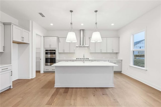 kitchen with a center island with sink, visible vents, wall chimney exhaust hood, light countertops, and pendant lighting