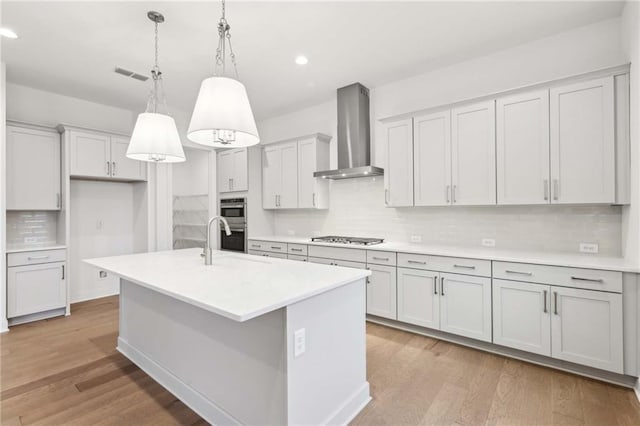 kitchen featuring visible vents, wall chimney exhaust hood, hanging light fixtures, a kitchen island with sink, and light countertops