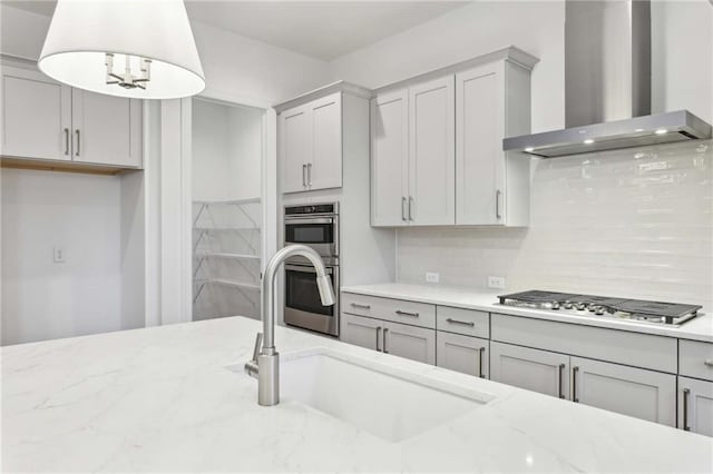 kitchen featuring stainless steel appliances, gray cabinetry, a sink, wall chimney range hood, and light stone countertops