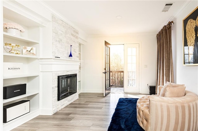 living room featuring built in shelves, a stone fireplace, ornamental molding, and light wood-type flooring