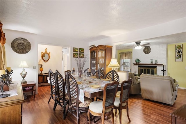 dining room with a brick fireplace, dark wood-type flooring, and ceiling fan