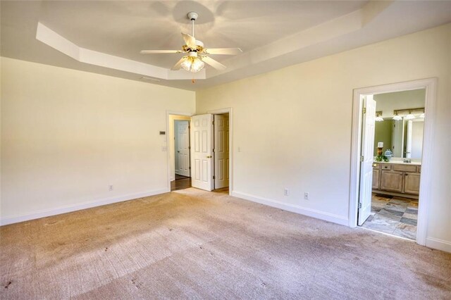 unfurnished bedroom with baseboards, ceiling fan, light colored carpet, a tray ceiling, and ensuite bathroom