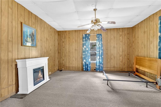 unfurnished room featuring a ceiling fan, a paneled ceiling, wood walls, a glass covered fireplace, and carpet flooring